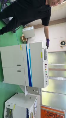 A person in black stands by a large, white industrial machine with control buttons and blue stripes, inside a workshop with green flooring, preparing to record a video in mp4 format for a Microsoft Teams presentation.