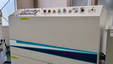 A large white industrial control panel with colored buttons, indicator lights, and switches on top, featuring blue diagonal stripes on the side. The panel is positioned indoors near a window, adjacent to a laptop used for video monitoring and HDD management.